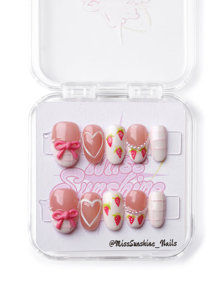 Kawaii pink press-on nails featuring strawberries, pearls, and heart details on a short oval shape.