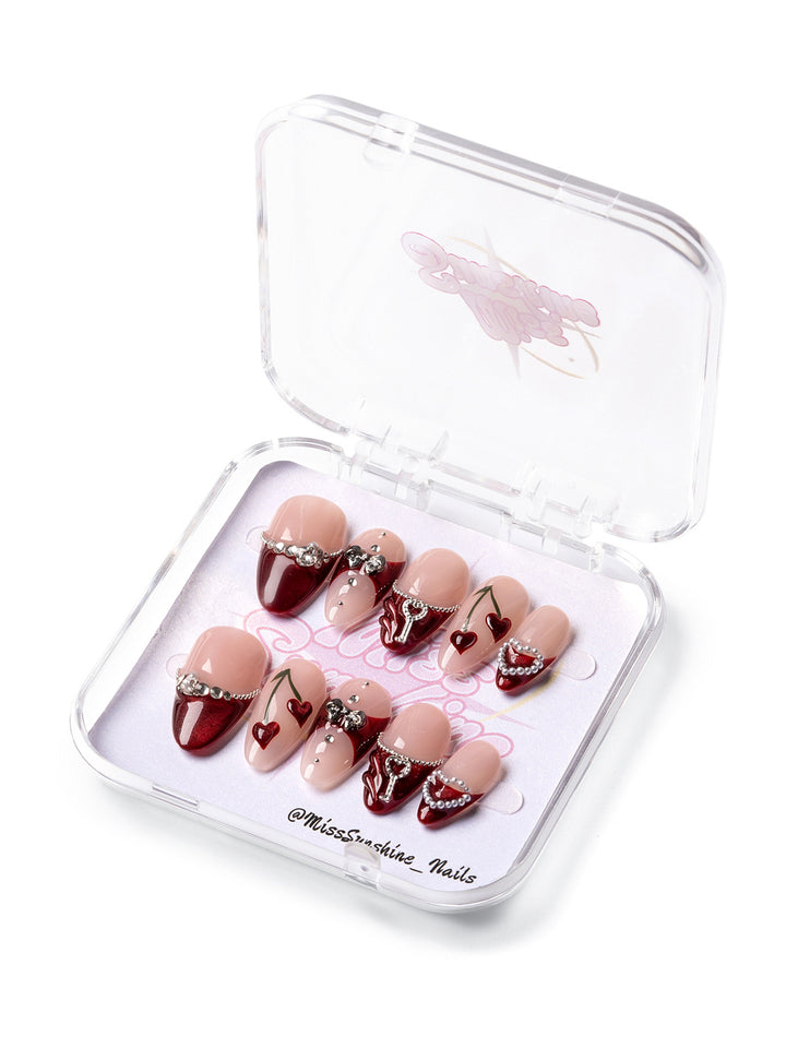 Cherry Kiss press-on nails featuring deep cherry red tips, nude tones, rhinestone chains, and heart accents for Valentine’s Day.